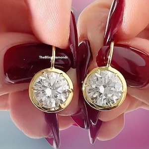 May include: A pair of gold earrings featuring large round clear diamonds. The earrings are held in the hands of a person with red nail polish.