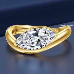 May include: A gold ring with a marquise-cut diamond. The diamond is set in a bezel setting, and the band has a smooth, curved design. The ring is displayed on a dark blue surface.