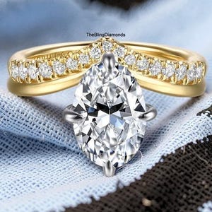 May include: A gold ring featuring a large, pear-shaped center stone and a V-shaped band adorned with a row of small, round, clear stones. The text TheBlingDiamonds is visible on the band. The ring has a polished finish and a prong setting.