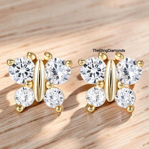 May include: A pair of gold butterfly-shaped stud earrings featuring four round clear stones each, arranged to form wings. The center of each butterfly is a smooth gold bar. The text TheBlingDiamonds is visible near the right earring.