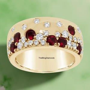 Puede incluir: Un anillo dorado ancho, con una banda adornada con piedras rojas y piedras redondas y claras alternas. El anillo tiene un acabado liso y pulido. El texto "TheBlingDiamonds" es visible en el interior de la banda.