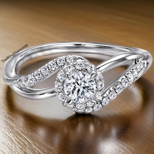 Puede incluir: Un anillo de compromiso plateado con un diamante central de talla redonda rodeado por un halo de diamantes más pequeños. La banda presenta un diseño de vástago dividido con diamantes pequeños adicionales. El anillo se muestra sobre una superficie de madera. El texto TheBlingDiamonds es visible.