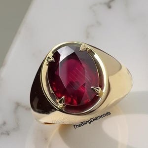 Pode incluir: Um anel de ouro com uma grande pedra preciosa vermelha escura de corte oval. A pedra preciosa é engastada em uma moldura de ouro com quatro garras. O anel tem uma banda lisa e polida. O texto "TheBlingDiamonds" é visível.
