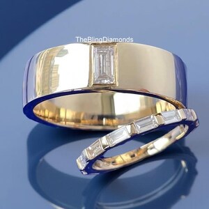 Puede incluir: Dos anillos de oro con detalles de diamantes. El anillo más grande presenta un solo diamante rectangular, mientras que el anillo más pequeño está adornado con múltiples diamantes rectangulares. Fondo azul.