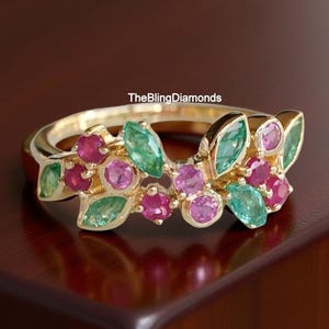 May include: A gold ring featuring a floral design with green, pink, and red gemstones. The ring has a band and is adorned with leaf-shaped green stones and round pink and red stones. The text "TheBlingDiamonds" is visible.