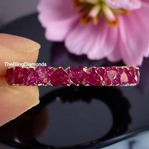 Könnte beinhalten: Ein goldener Ring mit herzförmigen, tiefroten Edelsteinen. Der Ring ist vor einem verschwommenen Hintergrund mit einer rosa Blume und einer dunklen Oberfläche platziert. Der Text "TheBlingDiamonds" ist sichtbar.