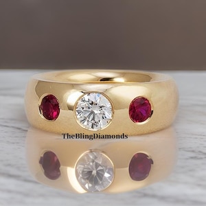 May include: A gold ring with a wide band, featuring a central clear diamond and two smaller red gemstones. The ring is highly polished, reflecting the light and the surface below. The text "TheBlingDiamonds" is visible.