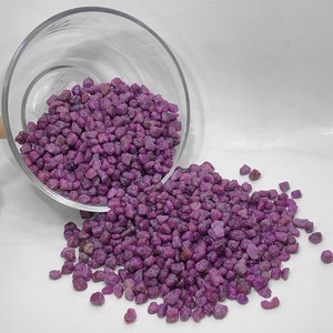 Könnte beinhalten: Ein klares Glasgefäß, das umgekippt ist und eine große Menge kleiner, unregelmäßig geformter, tiefmagentafarbener Steine ​​verschüttet. Die Steine ​​sind auf einer weißen Oberfläche verstreut und erzeugen einen auffälligen Kontrast.