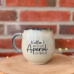 Könnte beinhalten: Cremefarbene Keramik-Tasse mit blauem Henkel und Rand. Die Tasse trägt den Text "Kaffee! Weil es für Aperol zu früh ist." in schwarzer Schrift. Die Tasse steht auf einer Holzoberfläche vor einem Ziegelhintergrund.