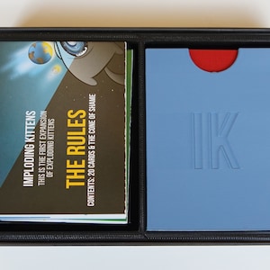 May include: A black box with a blue and red interior. The box contains a deck of cards with the text "Imploding Kittens" and "The Rules". The box also contains a blue card with the text "IK" in white.