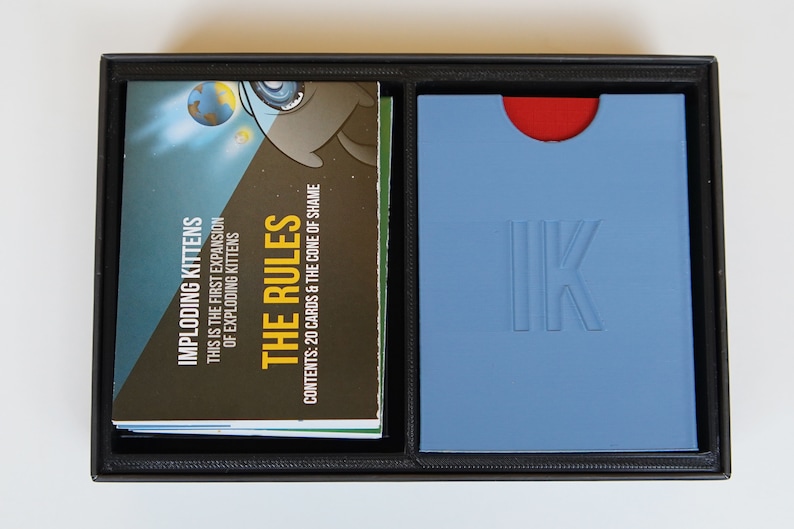 May include: A black box with a blue and red interior. The box contains a deck of cards with the text "Imploding Kittens" and "The Rules". The box also contains a blue card with the text "IK" in white.