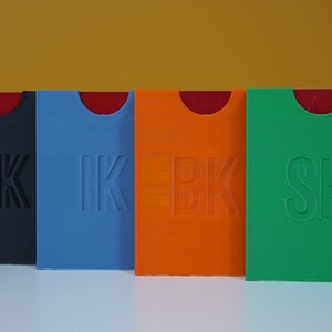 May include: Four small, rectangular boxes with rounded corners. The boxes are made of plastic and are in different colors: black, blue, orange, and green. Each box has a different letter combination embossed on the front: EK, IK, BK, and SK.