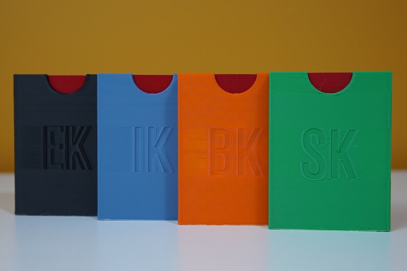 May include: Four small, rectangular boxes with rounded corners. The boxes are made of plastic and are in different colors: black, blue, orange, and green. Each box has a different letter combination embossed on the front: EK, IK, BK, and SK.