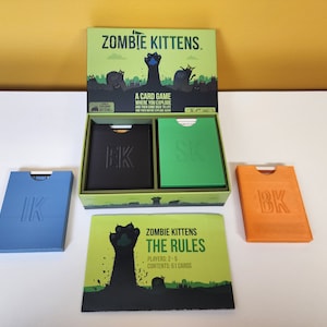 May include: An open box of the card game Zombie Kittens. The box is green with a black cat paw print on the front. The box contains two decks of cards, one black and one green, and a rule book. The rule book has a black cat paw print on the front and the text "Zombie Kittens The Rules". The text "Players: 2-5" and "Contents: 61 Cards" is also on the rule book.