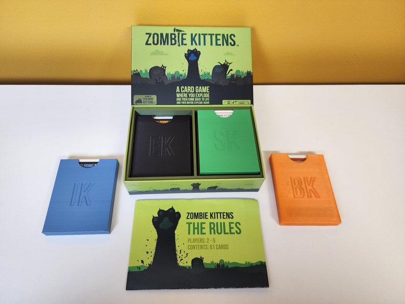 May include: An open box of the card game Zombie Kittens. The box is green with a black cat paw print on the front. The box contains two decks of cards, one black and one green, and a rule book. The rule book has a black cat paw print on the front and the text "Zombie Kittens The Rules". The text "Players: 2-5" and "Contents: 61 Cards" is also on the rule book.