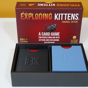 May include: A red box with the words "Exploding Kittens" and "Original Edition" printed on it. The box is open and shows two decks of cards, one black and one blue, with the letters "EK" printed on them.