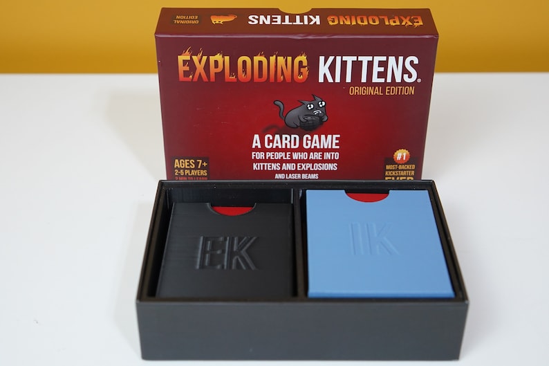 May include: A red box with the words "Exploding Kittens" and "Original Edition" printed on it. The box is open and shows two decks of cards, one black and one blue, with the letters "EK" printed on them.