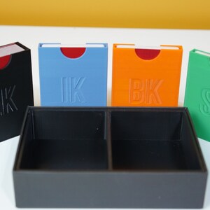May include: Four small boxes with red tops and different colored bases. The boxes are labeled "HK", "IK", "BK", and "SK". The boxes are sitting on a black tray with two compartments.