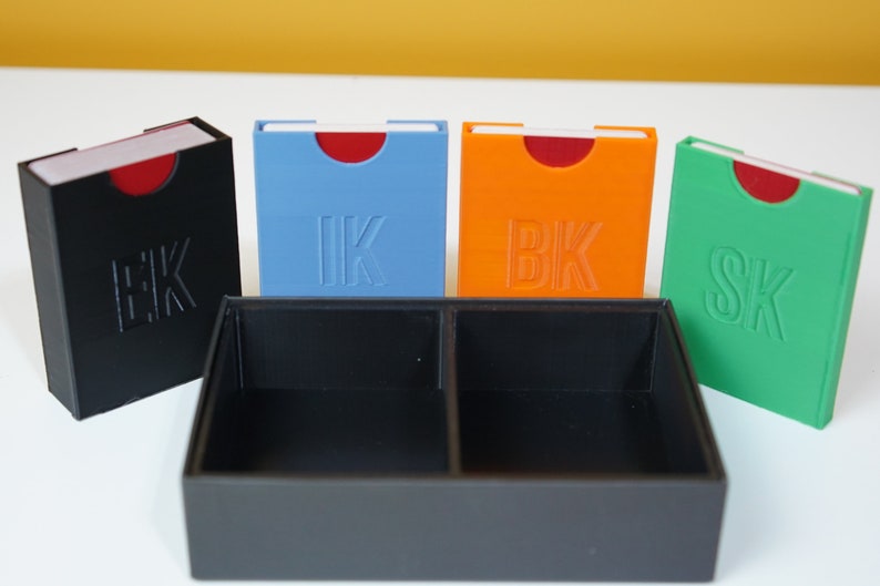 May include: Four small boxes with red tops and different colored bases. The boxes are labeled "HK", "IK", "BK", and "SK". The boxes are sitting on a black tray with two compartments.