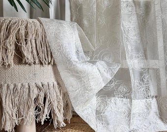 Könnte beinhalten: Transparente weiße Vorhänge mit zartem Rosenmuster. Eine gefranste Decke in einem neutralen Ton liegt auf einem Holzschemel. Die Szene wird von natürlichem Licht beleuchtet und erzeugt eine weiche, luftige Atmosphäre.