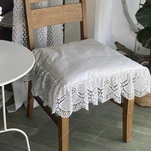 May include: A white chair cushion with a lace trim is placed on a wooden chair. The cushion has a square shape and a ruffled lace edge. The chair is made of light-colored wood.