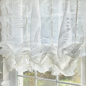 Puede incluir: Cortinas blancas y transparentes con un estampado de hojas de helecho. Las cortinas tienen volantes en la parte inferior y cuelgan frente a una ventana. Las hojas de helecho son de color gris claro.