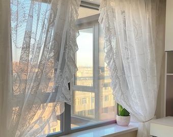 May include: White lace curtains with frills hang in a window with a view of a city skyline. A small potted plant sits on the windowsill.