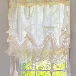 Op de afbeelding: Witte, doorschijnende, gerimpelde raambekleding met witte kwastbandjes. De bekleding is geplooid en gedrapeerd om een zachte, romantische uitstraling te creëren. De bekleding is opgehangen aan een raam met uitzicht op groene bomen.