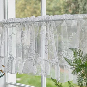 Puede incluir: Cortina de ventana de encaje blanco transparente con diseños florales y de hojas. La cortina tiene un borde superior e inferior con volantes, colgada de una barra plateada. La tela semitransparente permite el paso de la luz.