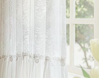 May include: White lace curtains with detailed floral and circular designs hang in front of a window. The curtains have a delicate, sheer appearance, with a ruffled bottom edge. The window frame is white, and the background shows a view of greenery.