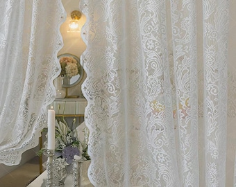 May include: White lace curtains with a scalloped edge and intricate floral patterns. The curtains are sheer, allowing light to filter through. A lit candle and a teacup are visible in the foreground, suggesting a cozy interior setting.