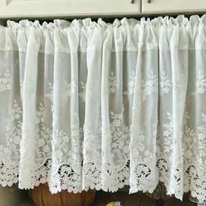 Könnte beinhalten: Ein weißer Scheibengardinen-Vorhang mit Stangendurchzug aus leichtem, transparentem Stoff. Der Vorhang ist mit einem komplexen weißen Blumenstickmuster verziert und schließt am unteren Rand mit einer bogenförmigen Spitzenkante ab.