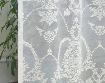 May include: White lace curtain featuring a detailed floral and geometric design. The curtain is semi-transparent, allowing light to pass through. The pattern includes stylized flowers, leaves, and circular shapes, creating a delicate and elegant look. The curtain's edge has a decorative border.