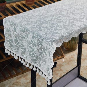 May include: A white lace table runner featuring a floral design and scalloped edging. The runner is decorated with small white tassels along the bottom. This delicate piece adds elegance to a table setting. (Approx. 150cm long)
