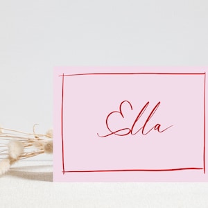May include: A pink card with a red border and the name "Ella" written in cursive with a heart shape above the first letter.