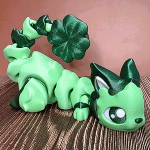 May include: A 3D-printed articulated cat-like figure in shades of green. The figure features a four-leaf clover design and a cat face with large eyes. The toy is designed to be flexible and poseable.