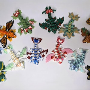 May include: A collection of twelve articulated dragon figurines in various colors and patterns, including butterfly-wing, mushroom, floral, and geometric designs. Each small creature features segmented bodies, detailed wings, and distinct color schemes.