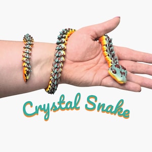 May include: A flexible articulated toy snake featuring a segmented body with a gradient of yellow, orange, and teal colors. The snake has a textured, crystalline appearance with dark brown accents on the scales and head. The text Crystal Snake is printed below.