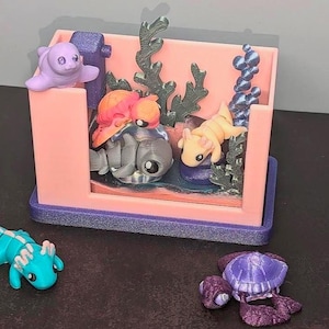 May include: A pink and purple miniature aquarium with a purple seahorse, a gray and purple seahorse, a pink and orange seahorse, a purple sea turtle, and a blue seahorse. The aquarium is filled with purple and green seaweed.