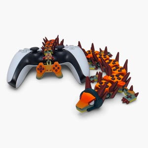 May include: A white and black video game controller featuring a custom orange and red articulated dragon-like attachment that extends from the top, with a detailed head piece wearing dark headphones and multiple spiked segments along its body.