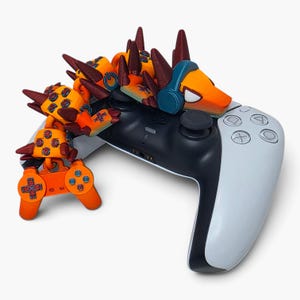 May include: A white and black video game controller with a 3D-printed orange and brown articulated creature figure draped over the top. The creature has dark brown spikes, blue headphones, and a power symbol on its back, with a small orange controller.
