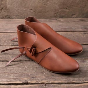 May include: A pair of ankle-high shoes crafted from smooth, medium-brown leather. The shoes feature a rounded toe, a wrap-around design that secures with a thin leather tie on the side, and a low-cut opening that exposes the ankle area.