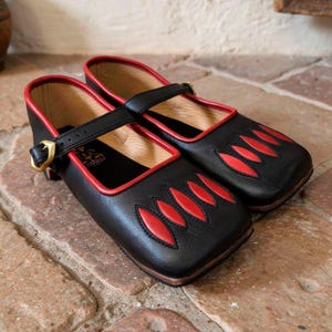 May include: A pair of black leather Mary Jane shoes featuring a square toe, red trim along the collar, and a decorative pattern of four red leaf-shaped cutouts on the vamp. Each shoe has a single adjustable strap with a gold-toned metal buckle.