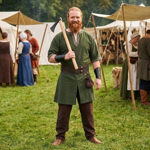 May include: A person with a red beard wearing a green tunic with a patterned neckline, brown trousers, a wide leather belt with a small pouch, and black leather wrist guards, holding a wooden-handled axe over their shoulder in a field.