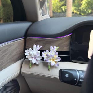 Könnte beinhalten: Zwei künstliche Blumenarrangements mit weißen, lavendelfarbenen und gelben Blütenblättern werden auf dem Armaturenbrett eines Autos präsentiert. Die Blumen haben grüne Stiele und sind in der Nähe der Bedienelemente des Autos platziert. Der Innenraum des Autos verfügt über Holzverkleidungen und ein cremefarbenes Armaturenbrett.