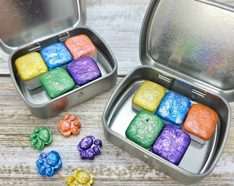 May include: Two silver metal tins with lids open, each containing six square watercolour paint pans. The pans are in shades of yellow, orange, green, blue, and purple with a shimmery finish. Six small paw print-shaped charms are scattered around the tins.