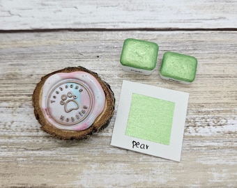 May include: A close-up of a green pear mica watercolor paint pan. The pan is labeled "pear" and is sitting on a wooden surface. There is a wax seal with a paw print and the text "Toocleens Kubbies" on the left side of the image.