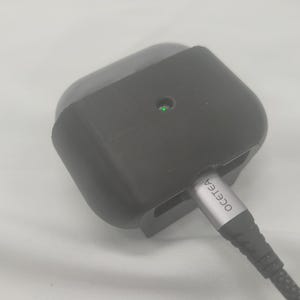 May include: A dark gray charging case for wireless earbuds. The case has a small green light and a charging cable plugged in. The cable has the brand name "OCETEA" printed on it.