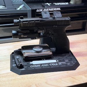 May include: A black handgun display stand featuring a textured brick-patterned base with the text "COME AND TAKE IT" in white lettering. The stand holds a black semi-automatic pistol with a mounted light, a folded pocket knife, and a brown leather wallet.