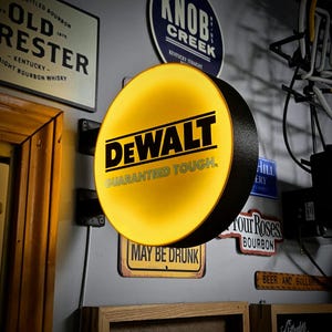 Może przedstawiać: Okrągły, podświetlany znak DeWalt z żółtą powierzchnią i czarnym napisem "DEWALT GUARANTEED TOUGH". Znak jest zamontowany na ścianie z innymi szyldami barowymi, w tym "Knob Creek" i "May Be Drunk".